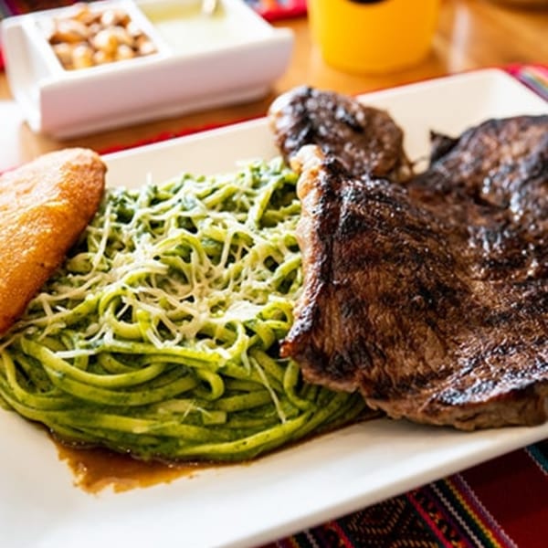 Peruvian pesto pasta with grilled steak and a golden fried plantain on a white plate.