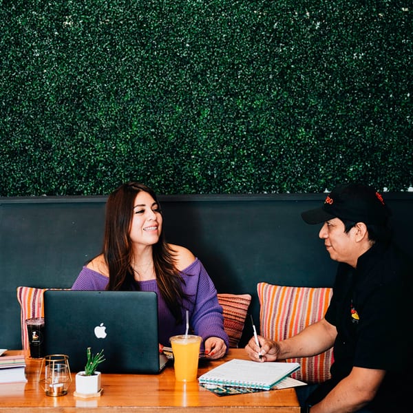 Two people, one with a laptop and one writing, at a wooden table with drinks and plants.