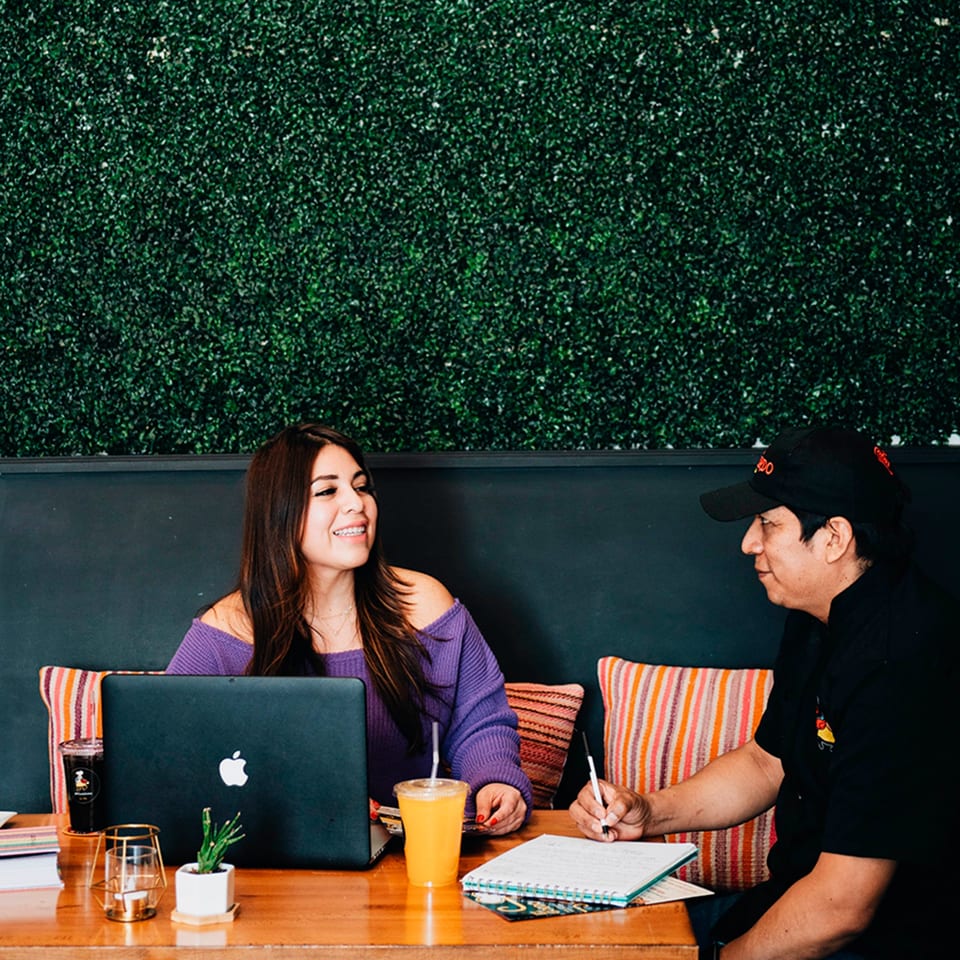 Two people, one with a laptop and one writing, at a wooden table with drinks and plants.