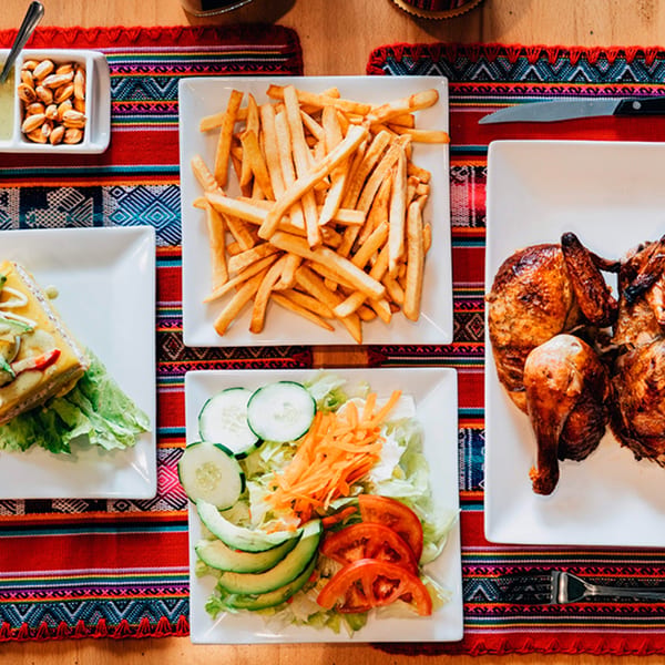 Peruvian meal with rotisserie chicken, french fries, salad, and a causa on a colorful tablecloth.