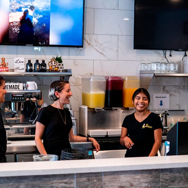 Two smiling El Gordo Peruvian Eatery employees stand behind a counter with juice dispensers and a coffee machine.