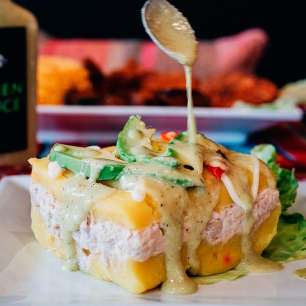 Peruvian causa rellena with avocado, red pepper, and creamy green sauce drizzled from a spoon.