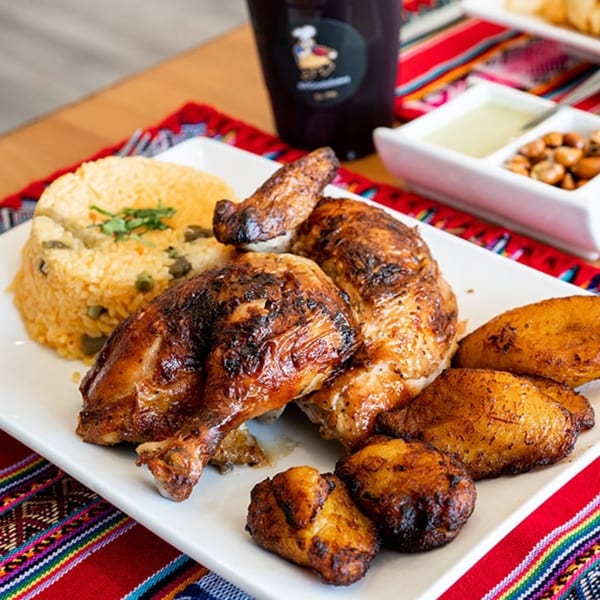 Peruvian rotisserie chicken with rice, fried plantains, and sauces on a colorful patterned tablecloth.