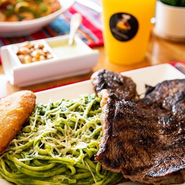 Peruvian steak with green pesto pasta, fried plantain, and a yellow drink on a colorful tablecloth.