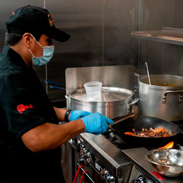 Chef in a black uniform, mask, and blue gloves stir-frying Peruvian food in a wok.