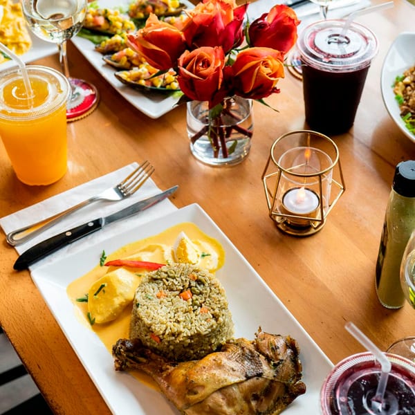 Peruvian meal with roasted chicken, rice, yellow sauce, drinks, and a rose centerpiece on a wooden table.