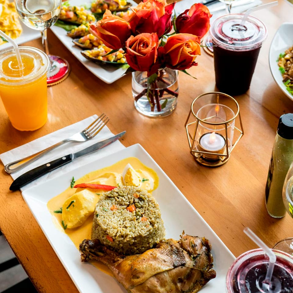 Peruvian meal with roasted chicken, rice, yellow sauce, drinks, and a rose centerpiece on a wooden table.